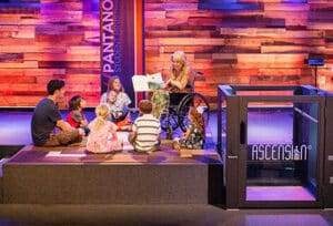 A teacher in a wheelchair reads to young students on a raised stage. A Protege wheelchair lift sits to the right side of the stage