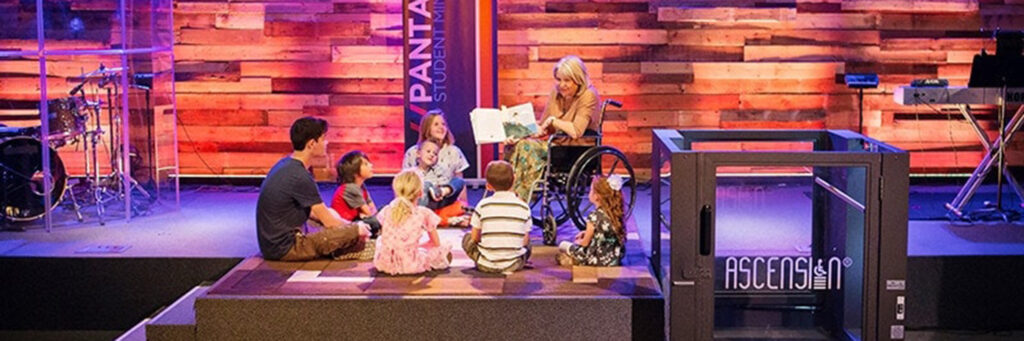 A teacher in a wheelchair reads to young students on a raised stage. A Protege wheelchair lift sits to the right side of the stage