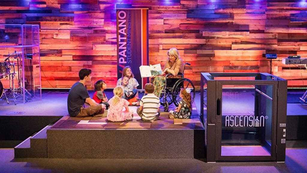 A teacher in a wheelchair reads to young students on a raised stage. A Protege wheelchair lift sits to the right side of the stage