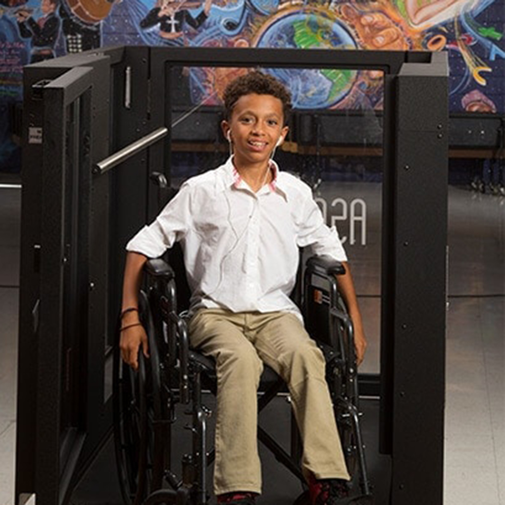 A black boy sits in a wheelchair in front of a Protege wheelchair lift