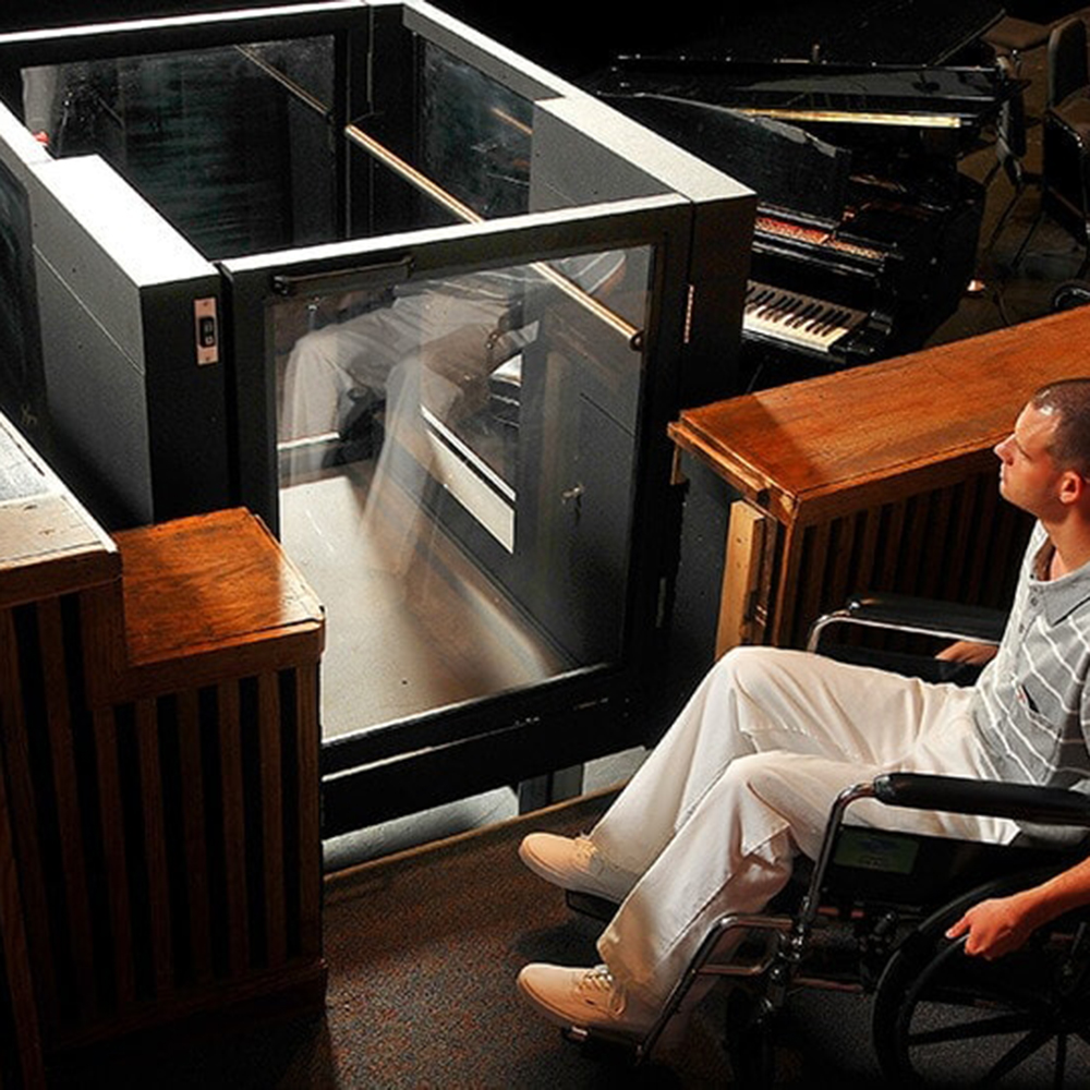 A white man sits in a wheelchair in front of a Protege wheelchair lift