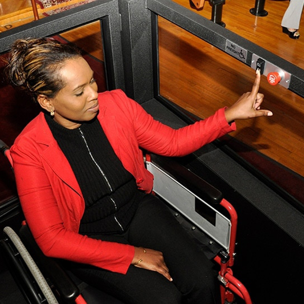 A black woman sits in a wheelchair inside a Virtuoso wheelchair lift